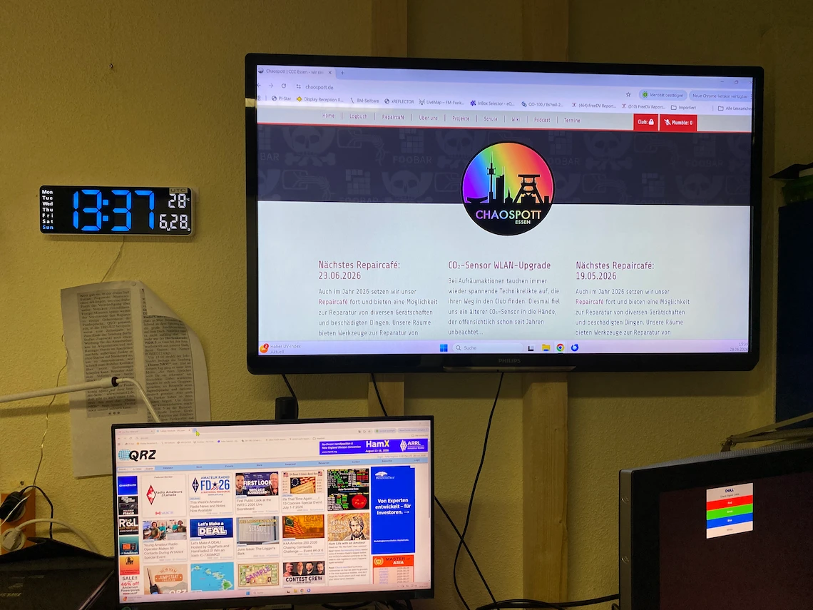 Innenaufnahme OV-Heim Romeo12. Ein großer an der Wand montierter Monitor zeigt die Website des Chaospott mit dem Vereinslogo und aktuellen Beiträgen. Darunter befindet sich ein weiterer Monitor mit der Amateurfunkplattform QRZ.com. Links an der Wand hängt eine digitale Uhr mit blauer LED-Anzeige (13:37 Uhr UTC).
